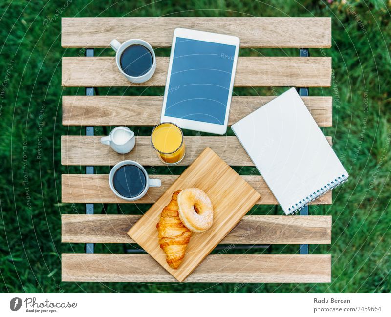 Frühstück im Grünen Garten mit französischem Croissant, Donuts, Kaffeetasse, Orangensaft, Tablette und Notizen Buch auf Holztisch Tisch Hintergrundbild Morgen