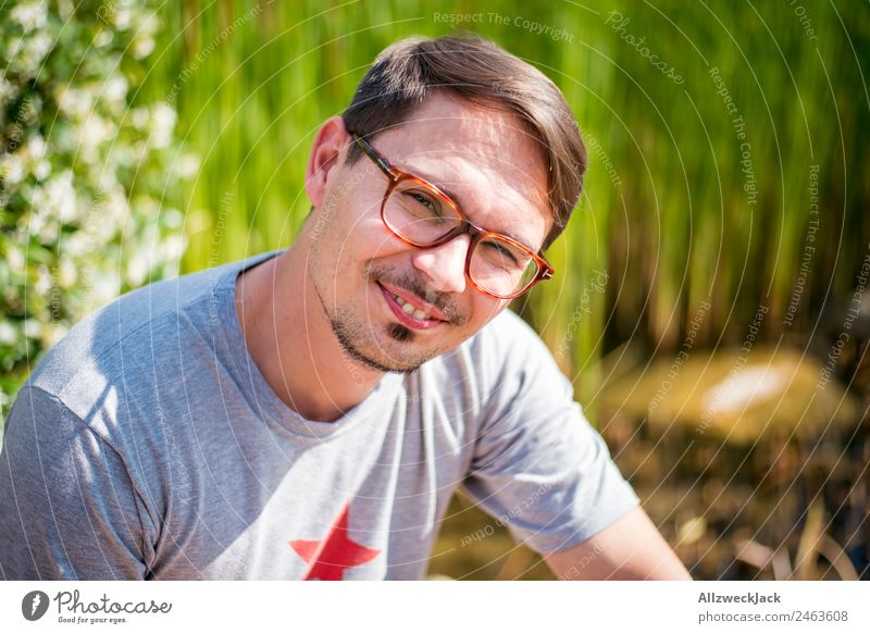 junger Mann mit Brille lächelt in Kamera 1 Mensch Junger Mann Porträt Brillenträger Lächeln sympathisch Fröhlichkeit Freundlichkeit Natur grün Schönes Wetter
