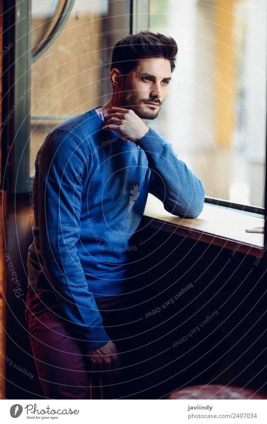 Nachdenklicher Mann im blauen Pullover mit verlorenem Blick am Fenster in einer modernen Kneipe. Bärtiger Mann mit moderner Frisur Lifestyle Stil schön