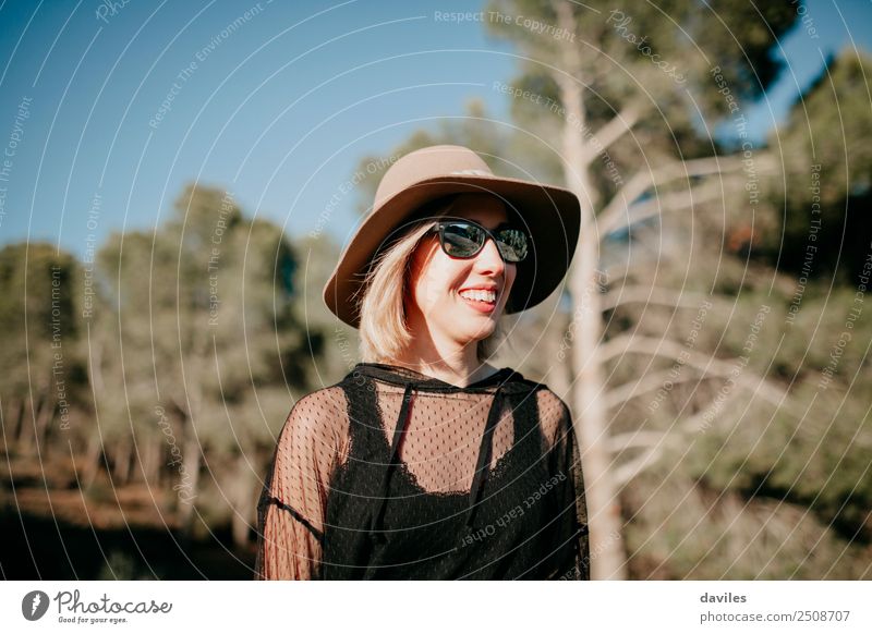 Hübsches blondes Mädchen mit Sonnenbrille und Hut, das sich in der Natur mit einem Kiefernwald im Hintergrund amüsiert Lifestyle Stil Freude Erholung