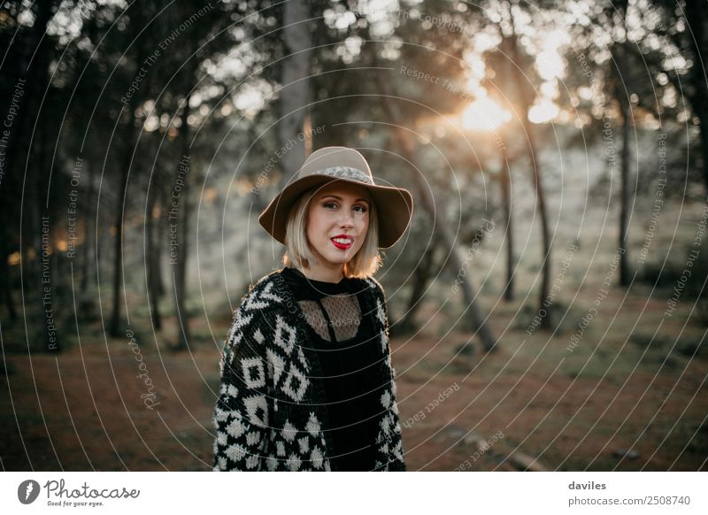 Blonde Frau mit Hut, die bei Sonnenuntergang mit der Sonne im Hintergrund im Wald spazieren geht. Lifestyle Freude schön Ferien & Urlaub & Reisen Tourismus