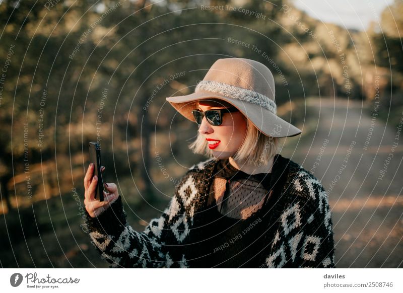 Blonde junge Frau mit Hut und Sonnenbrille, die bei Sonnenuntergang im Wald mit einem Mobiltelefon ein Foto macht. Lifestyle elegant Stil Erholung Telefon