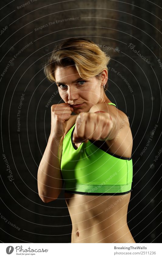 Nahaufnahme Frontalporträt einer jungen sportlichen Frau in Sportkleidung in einer Turnhalle vor dunklem Hintergrund, stehend in Boxhaltung mit Händen und Fäusten, Blick in die Kamera