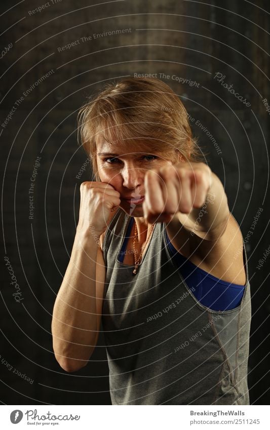 Nahaufnahme Frontalporträt einer jungen sportlichen Frau mittleren Alters in Sportkleidung im Fitnessstudio vor dunklem Hintergrund, stehend in Boxhaltung mit Händen und Fäusten, in die Kamera blickend