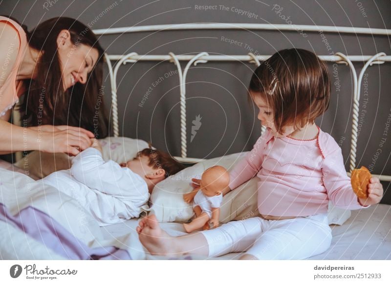 Glückliches kleines Mädchen hält Puppe und Keks, während Frau spielt mit einem Jungen über dem Bett. Weekend Familie Freizeit Konzept. Essen Frühstück Lifestyle