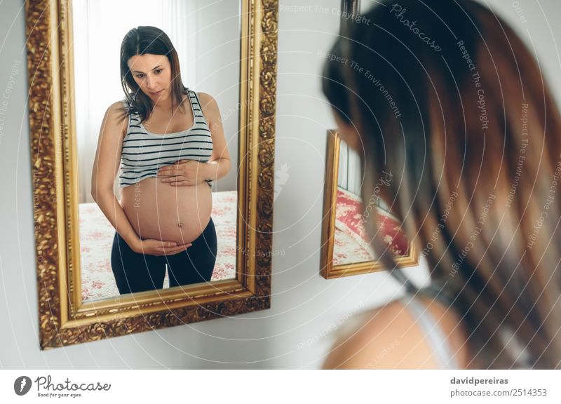 Schwangere Frau, die ihren Bauch ansieht. Lifestyle schön Leben Spiegel Schlafzimmer Mensch Baby Erwachsene Eltern Mutter Hand Denken Wachstum warten