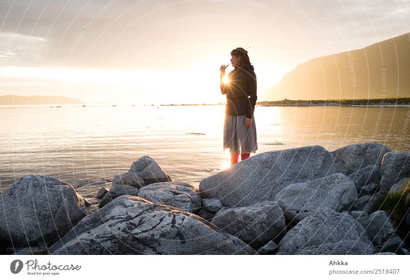 Junge Frau mit Pfeife steht am Ufer des Nordmeeres auf Steinen vor der Mitternachtssonne Gegenlicht Sonne ufer Fjord Felsen Meer Licht durchscheinend erhaben