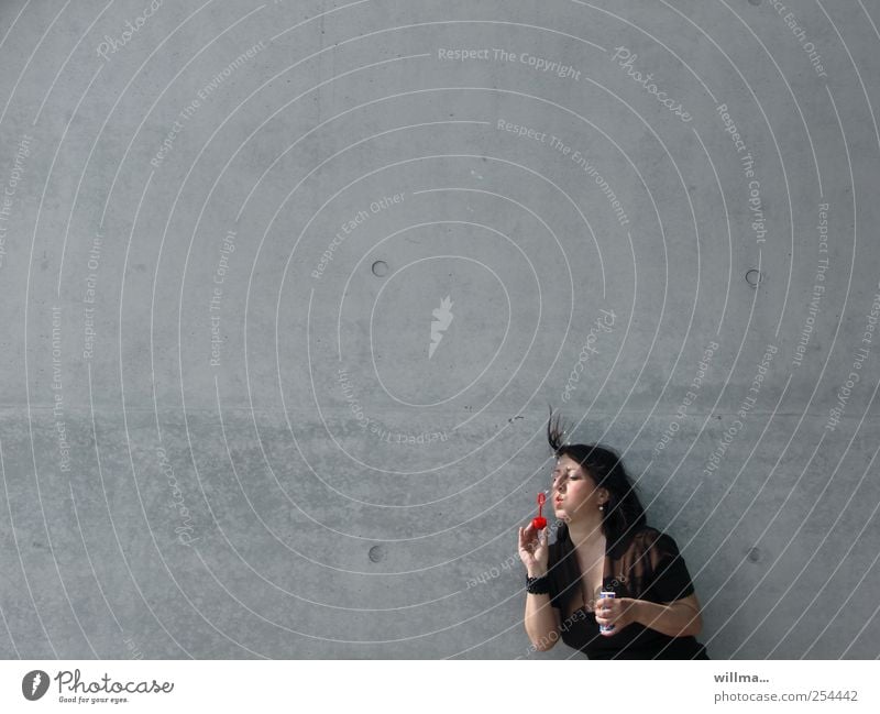 Frau bläst mit Hingabe Seifenblasen vor einer grauen Wand, neutraler Hintergrund Erwachsene 1 Mensch Mauer brünett Spielen Leichtigkeit einzeln Betonwand