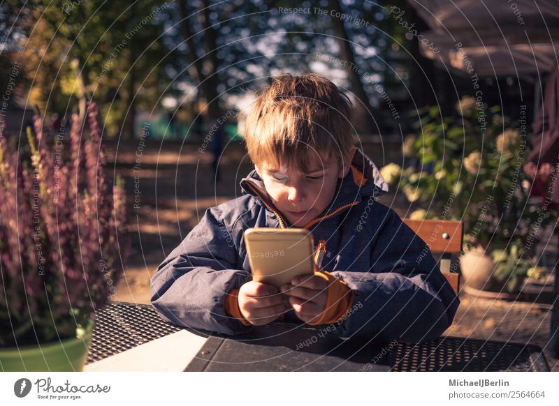 Junge sitzt mit Smartphone an einem Tisch im Freien Mensch maskulin Kind Kindheit 1 3-8 Jahre Herbst Spielen Jacke herbstlich kalt frisch Außenaufnahme PDA