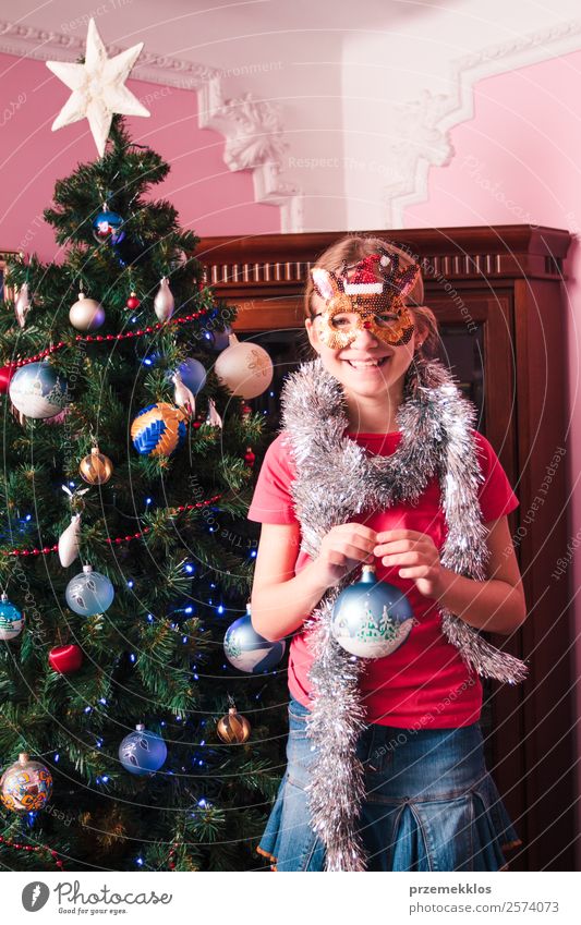 Junges Mädchen schmückt Weihnachtsbaum, hält große Weihnachtskugel. Junges blondes Mädchen in blauem Jeansrock, rosa Bluse und Strumpfhose. Mädchen hat eine lustige Rentiermaske auf dem Gesicht. Junges Mädchen feiert Weihnachten zu Hause