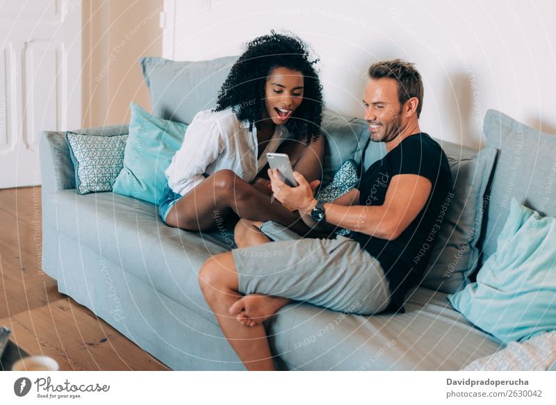 Ein glückliches junges Paar entspannte sich zu Hause auf der Couch, las ein Buch und schaute auf das Handy. interrassisch Partnerschaft Jugendliche lesen