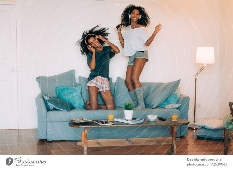 Glückliche junge zwei schwarze Frauen sitzen und stehen auf der Couch am Computer und Handy mit tanzenden Kopfhörern. Mobile Telefon hören nach oben Tanzen