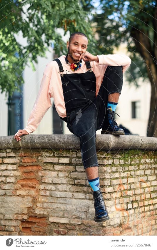 Junger schwarzer Mann in Freizeitkleidung und mit Kopfhörern sitzt vor einem städtischen Hintergrund. Lifestyle Glück schön Mensch maskulin Junger Mann