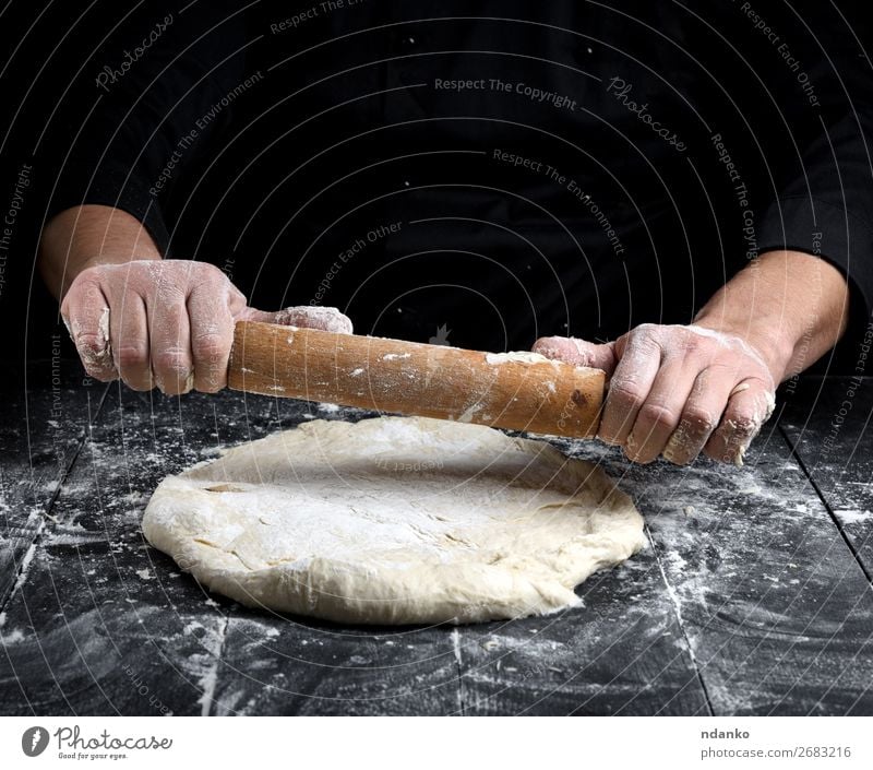 Chefkoch in einer schwarzen Tunika rollt einen Teig für eine runde Pizza. Teigwaren Backwaren Brot Ernährung Tisch Küche Koch Mann Erwachsene Hand Holz machen