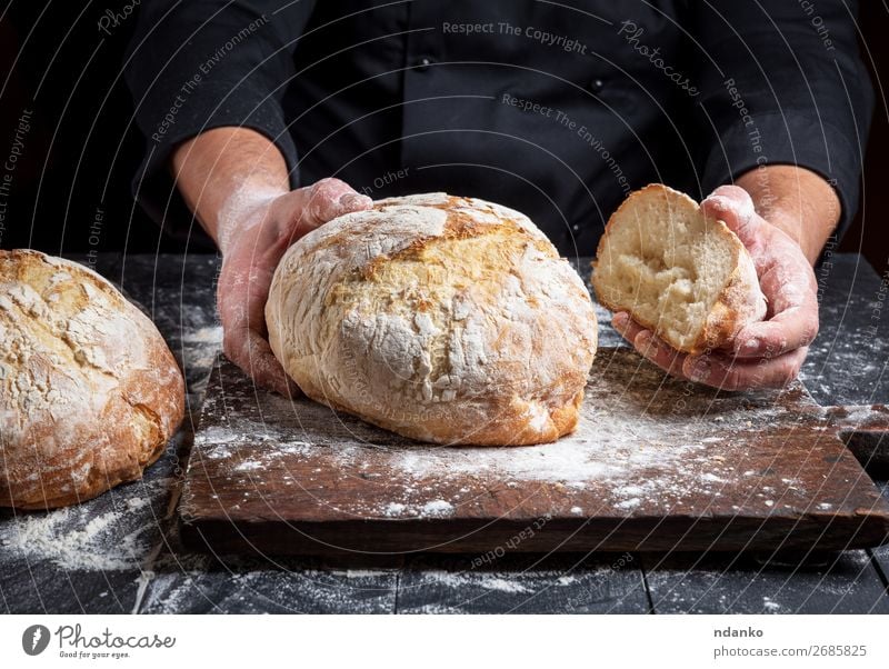 Koch in einer schwarzen Tunika hält frisch gebackenes Brot bereit. Ernährung Tisch Küche Mensch Hand Finger Holz machen dunkel braun weiß Tradition Bäcker