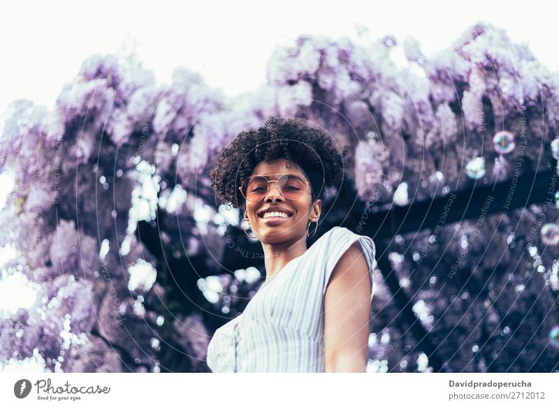 Fröhliche junge schwarze Frau, umgeben von Blumen. Blüte Frühling Fliederbusch Porträt multiethnisch Afrikanisch Person gemischter Abstammung Lächeln