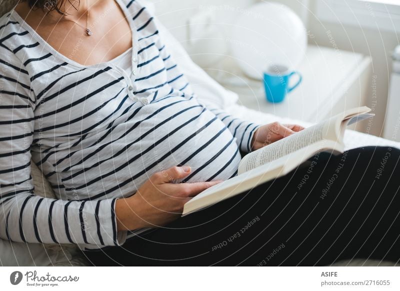 Glückliche schwangere Frau im gestreiften T-Shirt sitzt auf dem Bett mit den Beinen auf einem Kissen und liest Bücher über Schwangerschaft, Nahaufnahme schön