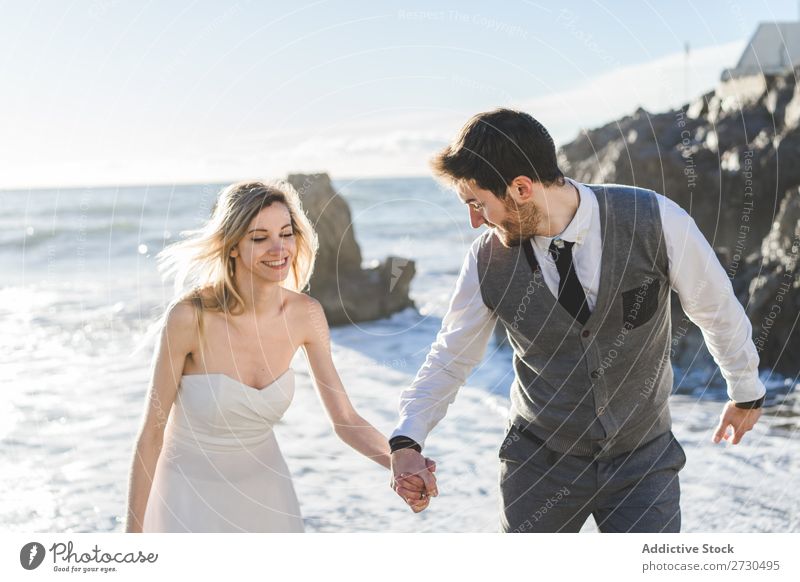 Schönes Brautpaar, das am Ufer spazieren geht. Paar hochzeitlich Strand Fröhlichkeit Seeküste Händchenhalten heiter Außenaufnahme Zusammensein Zufriedenheit