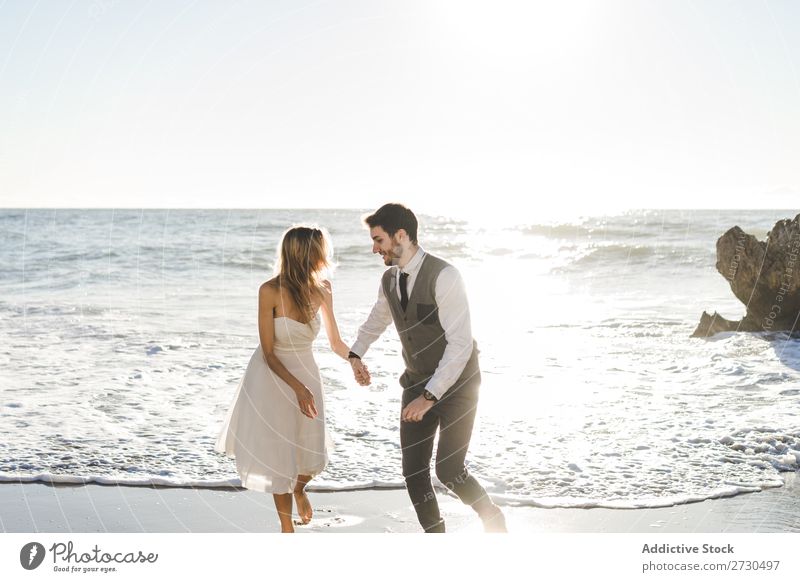 Schönes Brautpaar, das am Ufer läuft. Paar hochzeitlich rennen Strand Fröhlichkeit Seeküste Händchenhalten heiter Außenaufnahme Zusammensein Zufriedenheit