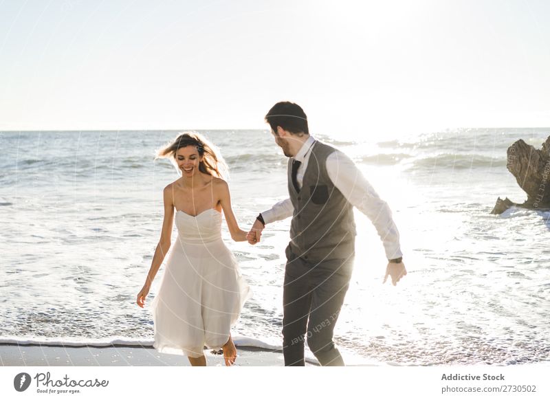 Schönes Brautpaar, das am Ufer läuft. Paar hochzeitlich rennen Strand Fröhlichkeit Seeküste Händchenhalten heiter Außenaufnahme Zusammensein Zufriedenheit