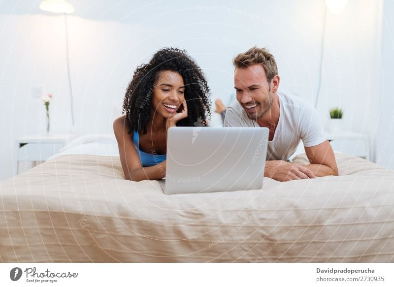 Ein glückliches junges Paar entspannte sich zu Hause und lag am Computer im Bett. interrassisch Partnerschaft Jugendliche Schlafzimmer Technik & Technologie