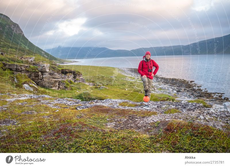 Junger Mann mit Fernglas posiert an einem Fjord im Abendlicht mit Blick auf Meer und Berge rot posieren grün Stolz forschen Neugier Freiheit Menschenleer