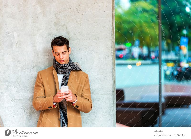 Vorderansicht eines modischen jungen Mannes in Jeanskleidung, der sich an eine Wand lehnt, während er im Freien ein Mobiltelefon benutzt Lifestyle elegant Stil