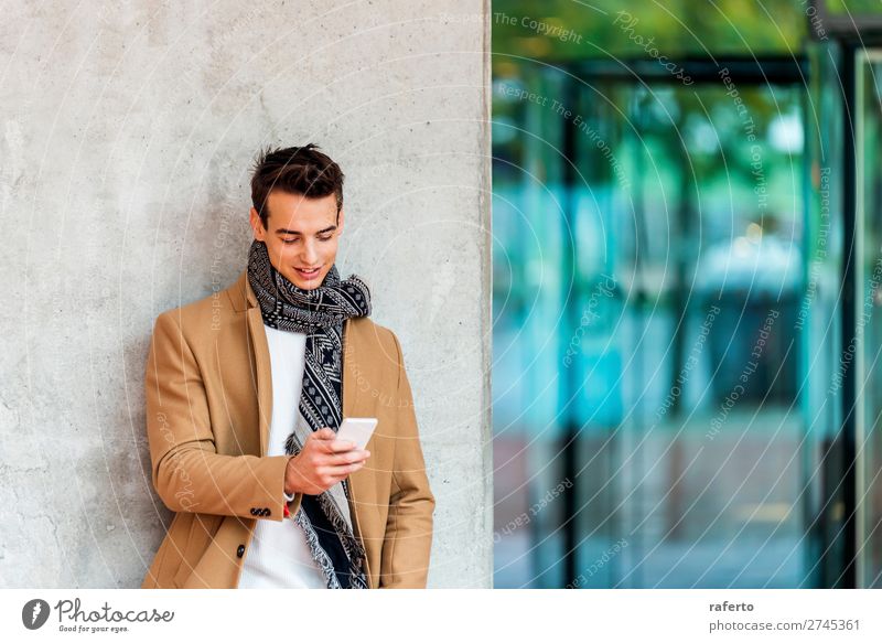 Vorderansicht eines modischen jungen Mannes in Jeanskleidung, der sich an eine Wand lehnt, während er im Freien ein Mobiltelefon benutzt Lifestyle elegant Stil