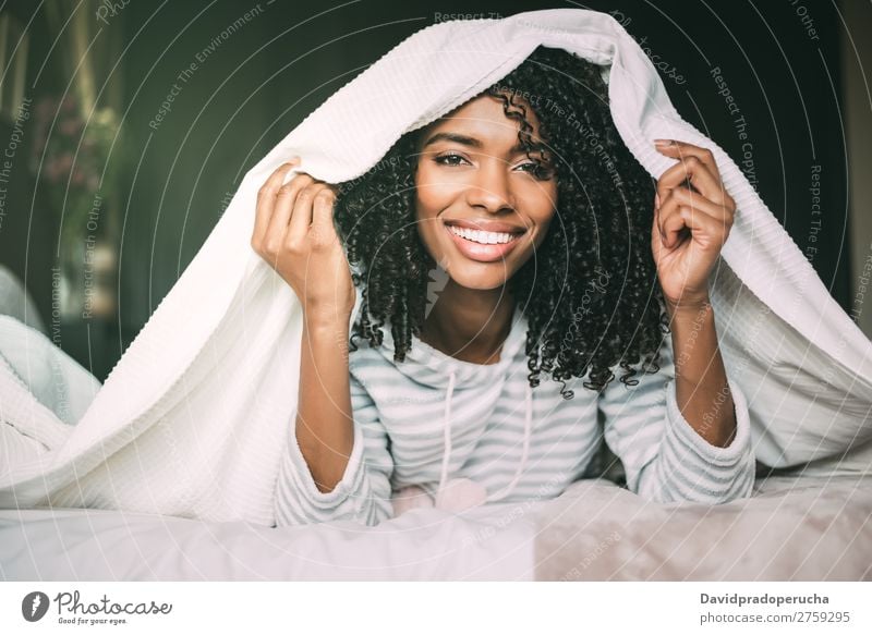 Nahaufnahme einer hübschen schwarzen Frau mit lockigem Haar, die lächelt und mit Bettwäsche auf dem Bett bedeckt ist, mit Blick auf die Kamera. schwarze Frau