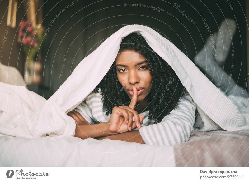 attraktive schwarze Frau, die um Stille bittet, mit dem Finger auf den Lippen auf dem Bett. schön Gesicht geheimnisvoll gestikulieren Porträt Nahaufnahme lügen