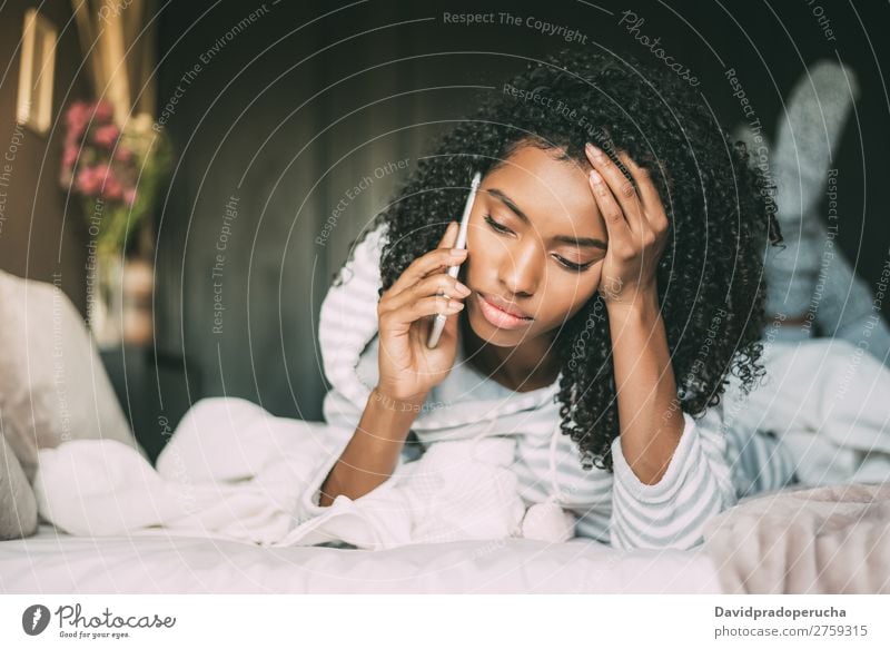 Wunderschöne, ernsthaft nachdenkliche und traurige schwarze Frau mit lockigen Haaren mit Smartphone auf dem Bett. Mobile Telefon PDA herabsehend Traurigkeit Wut