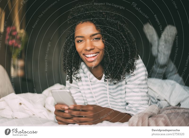 Nahaufnahme einer hübschen schwarzen Frau mit lockigem Haar und Smartphone auf dem Bett mit Blick auf die Kamera. schwarze Frau PDA Mobile Telefon Porträt lügen