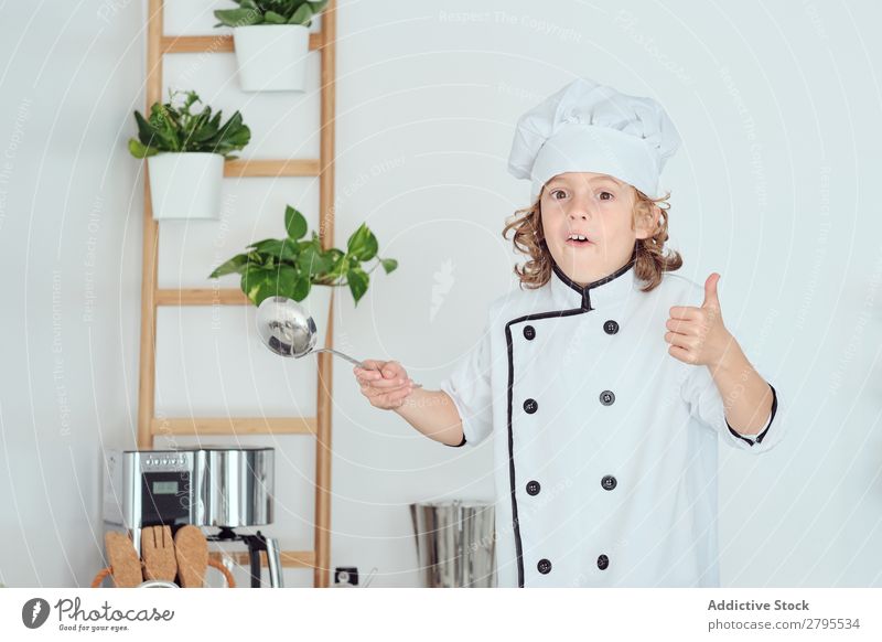 Junge mit Kochmütze, der die Schöpfkelle hält und den Daumen in der Küche zeigt. Schöpflöffel Daumen hoch Küchenchef Kind Hut Coolness gestikulieren
