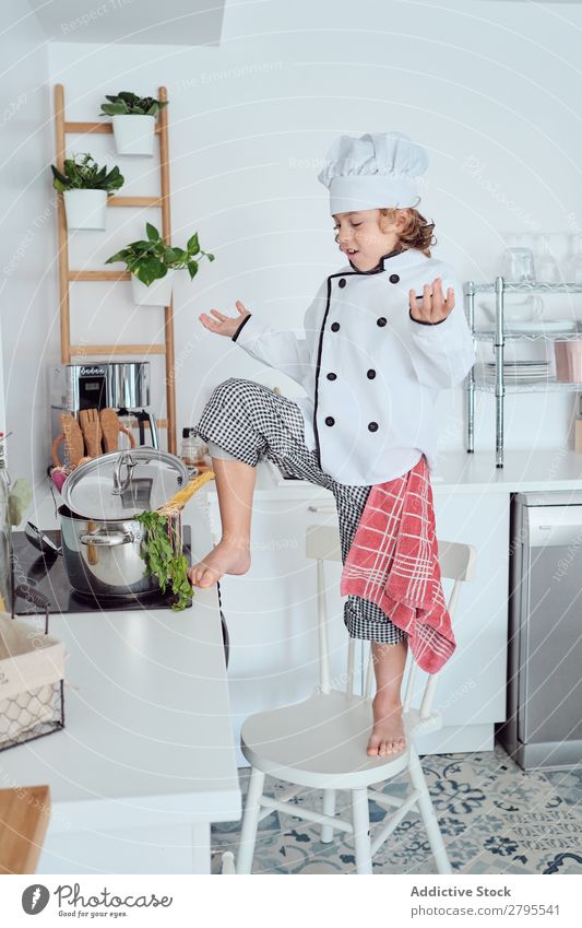 Junge mit Kochmütze mit erhobener Hand in der Nähe des Topfes auf Stuhl in der Küche. erhöht Küchenchef Kind Gemüse Hut kochen & garen modern lustig heimwärts