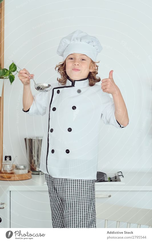 Junge mit Kochmütze hält Schöpfkelle in der Nähe des Mundes in der Küche. Schöpflöffel Küchenchef Kind Topf Verkostung Lebensmittel Hut kochen & garen Metall