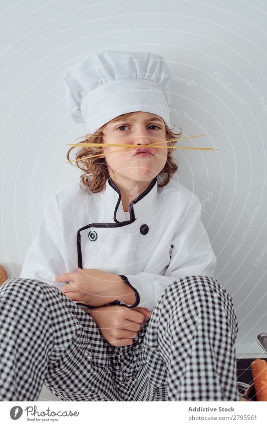 Junge mit Kochmütze, der in der Küche einen Schnurrbart aus Makkaroni macht. Oberlippenbart Küchenchef Kind Hut Spätzle regenarm machen kochen & garen modern