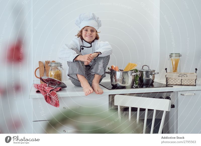 Junge sitzt in der Nähe von Töpfen auf der Elektrofritteuse in der Küche. Koch Topf Küchenchef Kind Gemüse Hut Herd & Backofen kochen & garen modern lustig