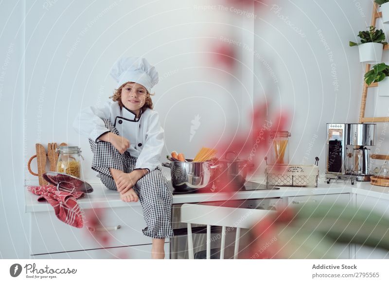 Junge sitzt in der Nähe von Töpfen auf der Elektrofritteuse in der Küche. Koch Topf Küchenchef Kind Gemüse Hut Herd & Backofen kochen & garen modern lustig