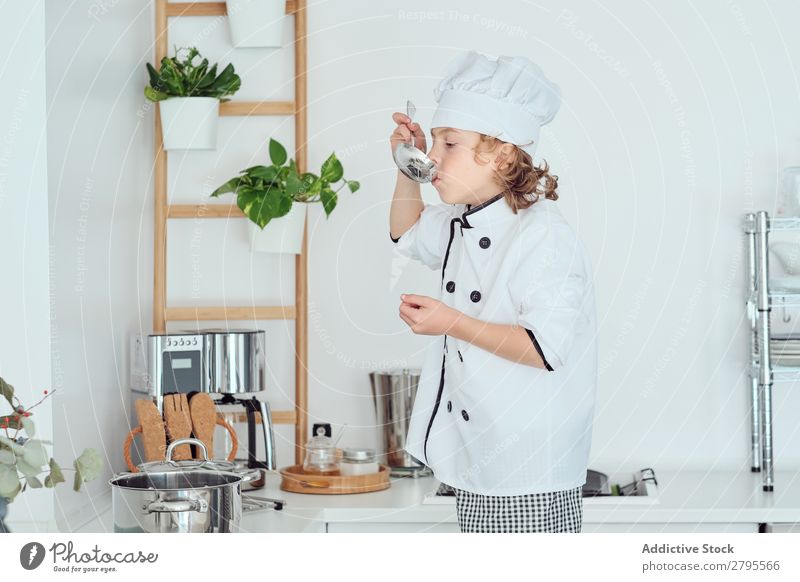 Junge mit Kochmütze hält Schöpfkelle in der Nähe des Mundes in der Küche. Schöpflöffel Küchenchef Kind Topf Verkostung Lebensmittel Hut kochen & garen Metall
