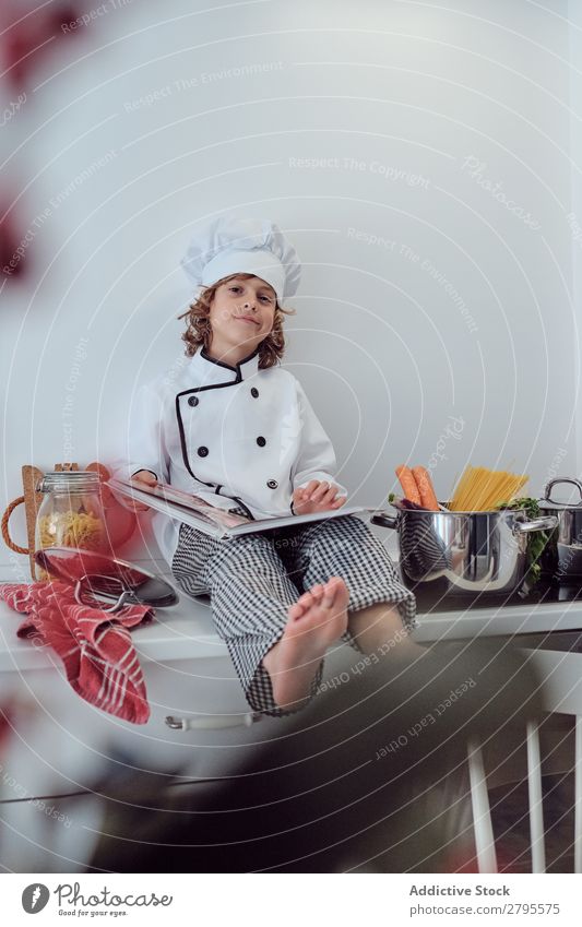 Junge mit Kochmütze und Buch in der Nähe von Töpfen auf der Elektrofritteuse in der Küche. Topf Küchenchef Kind Gemüse Hut Lautstärke lesen Herd & Backofen