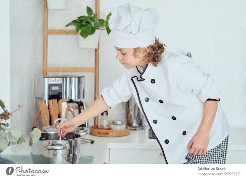 Junge mit Kochmütze mit erhobener Hand in der Nähe des Topfes auf Stuhl in der Küche. erhöht Küchenchef Kind Gemüse Hut kochen & garen modern lustig heimwärts