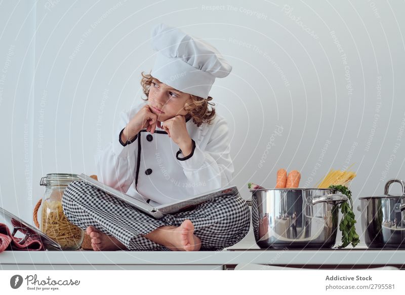Junge mit Kochmütze und Buch in der Nähe von Töpfen auf der Elektrofritteuse in der Küche. Topf Küchenchef Kind Gemüse Hut Lautstärke lesen Herd & Backofen