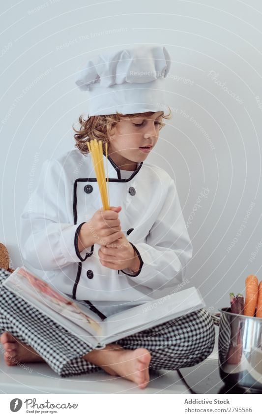Junge mit Kochmütze, der Nudeln in den Topf auf die Elektrofritteuse in der Küche legt. Spätzle Küchenchef Kind Makkaroni Möhre Gemüse Hut Herd & Backofen