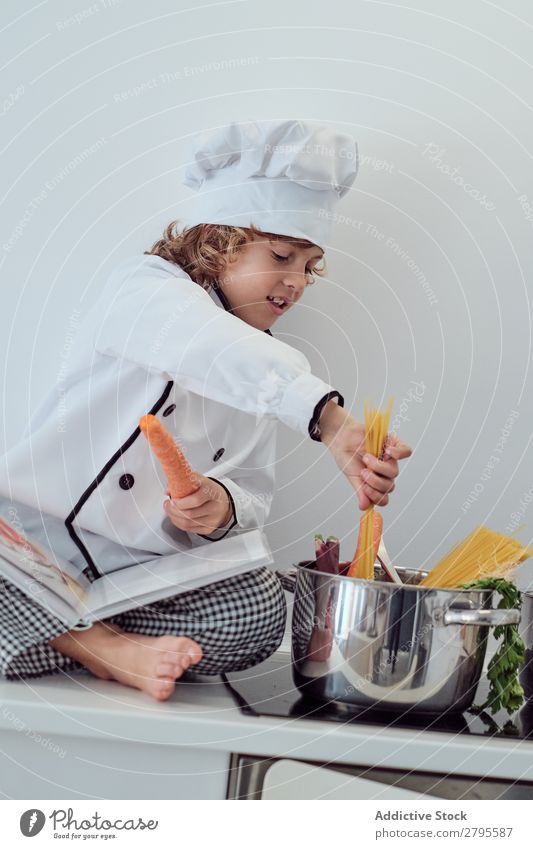 Junge mit Kochmütze, der Nudeln in den Topf auf die Elektrofritteuse in der Küche legt. Spätzle Küchenchef Kind Makkaroni Möhre Gemüse Hut Herd & Backofen