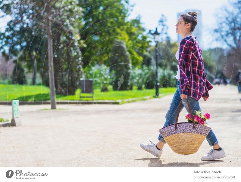 Seitenansicht einer jungen Hipsterfrau, die einen Weidenkorb hält, während sie bei Sonnenschein in einem Park spazieren geht. Profil Junge Frau Schickimicki