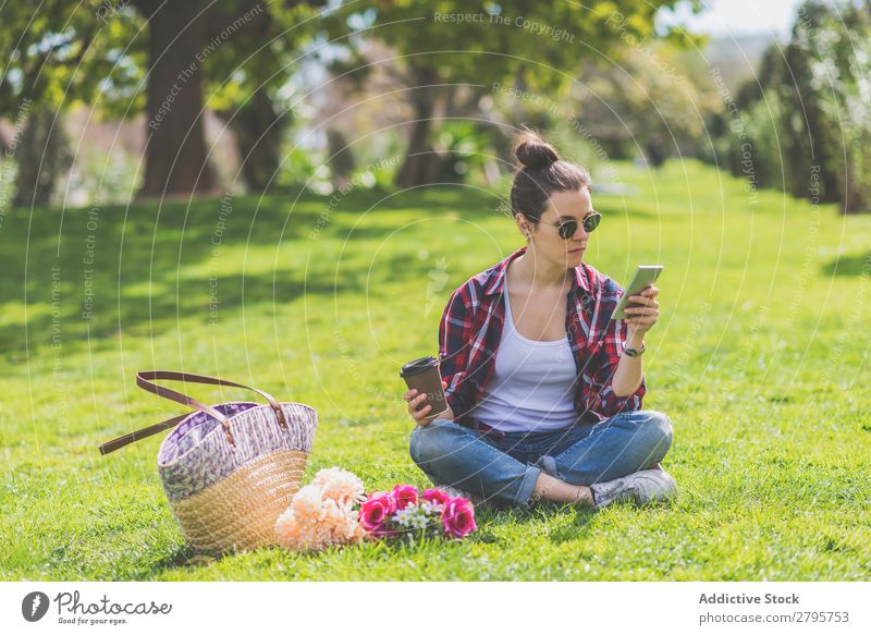 Vorderansicht einer jungen Hipsterfrau mit Sonnenbrille, die auf Gras in einem Park sitzt und dabei ein Handy benutzt. Junge Frau Schickimicki trendy sitzen