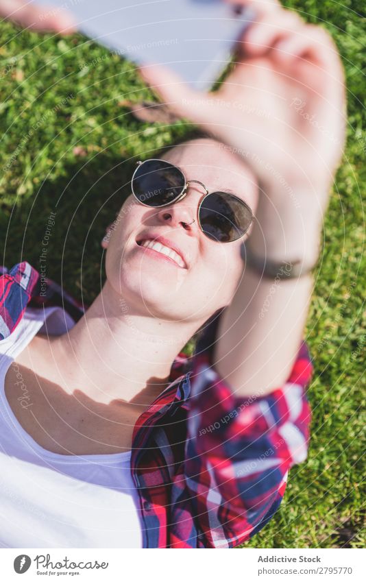 Oben die junge lächelnde Hipsterfrau, die an einem sonnigen Tag im Park auf Gras liegt und dabei mit dem Handy ein Selfie nimmt. obere Ansicht Junge Frau
