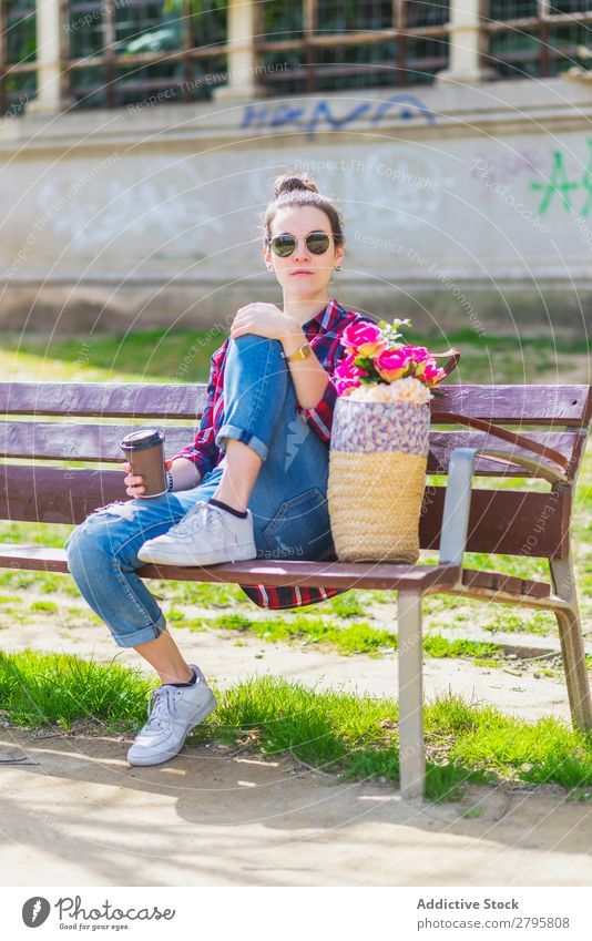 Frontansicht einer jungen Hipsterfrau, die auf einer Parkbank sitzt und sich an einem sonnigen Tag entspannt und in die Kamera schaut. Vorderansicht Junge Frau