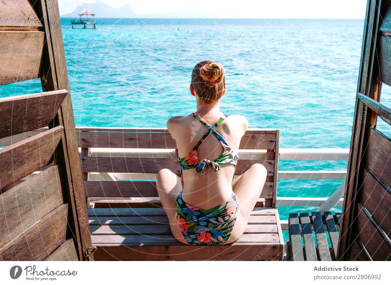 Frau im Bikini sitzt auf einer Bank in der Nähe des Wassers. Meer Jamaika Sonnenbrille Badeanzug Sitz blau tropisch exotisch sitzen Accessoire Sommer dünn Dame