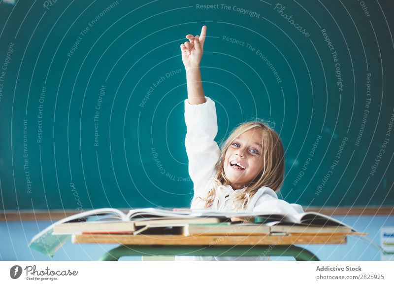 Fröhliches Schulmädchen mit erhobener Hand Mädchen Klassenraum Tafel sitzen Schreibtisch Lächeln Glück hochreichen Antwort niedlich Bildung Schule Schulklasse